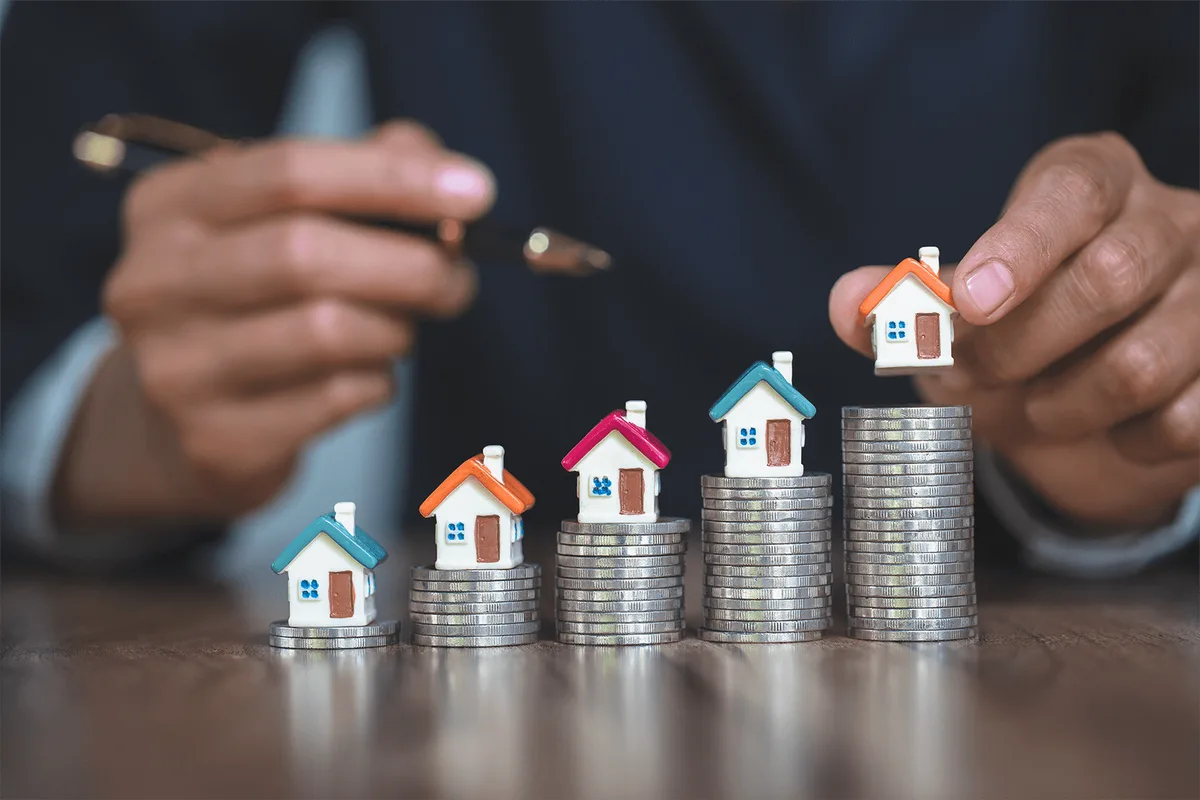 Miniature houses resting on rising stacks of coins while a hand places the tallest house on top